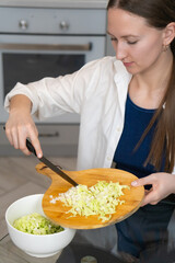 Woman wearing a white shirt sliding chopped cabbage from a wooden cutting board into a white bowl, surrounded by a modern kitchen setting, emphasizes healthy meal preparation. Vertical photo