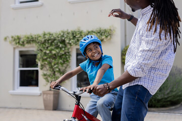 Obraz premium African American father teaching son to ride bicycle outside, both smiling happily