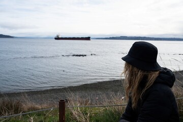 Obraz premium A woman in a black hat and coat sits on a grassy shoreline, gazing toward a distant cargo ship