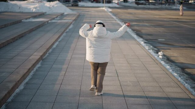 Back view of youth grooving down tiled walkway beside steps on busy winter street lined with snow piles, wearing puffy white jacket and beanie, surrounded by billboards, and city buildings