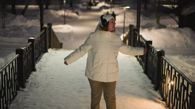Cheerful woman in white puffer coat and headphones dancing on snow covered pavilion stairs at night with faint silhouettes in background moving through illuminated winter park scene gracefully