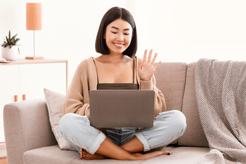 Online Video Call. Portrait of smiling asian lady making virtual conference on laptop, waving with hand to webcam. Communication Concept. Woman chatting using computer, sitting on the couch at home