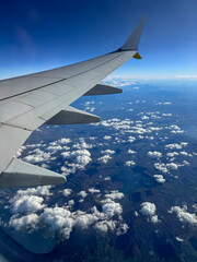 Obraz premium AERIAL: Stunning view from airplane window of a wing above scattered cloud layer and mountainous landscape. Traveling by aeroplane high above vast and rugged European terrain under a clear blue sky.