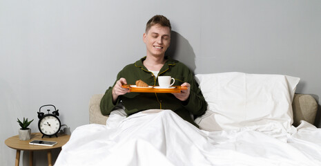 Happy man in his twenties holds a breakfast tray with a croissant and coffee. Peaceful indoor...