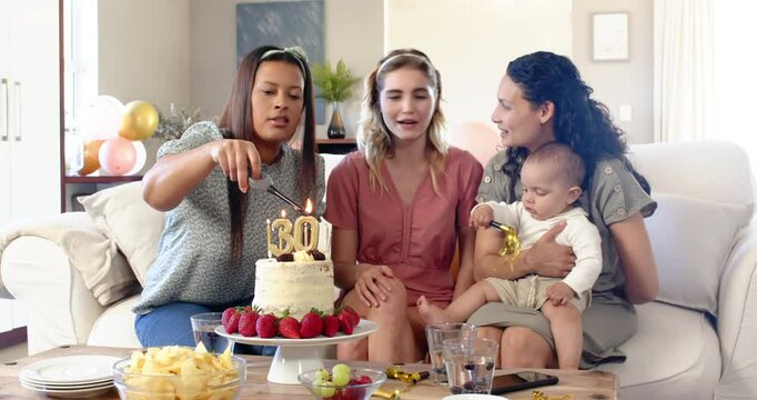Three happy multiracial and white female friends with baby blowing candles on birthday cake at a bir