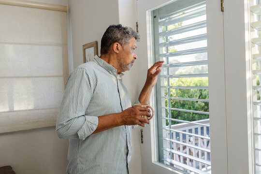Senior man holding coffee, looking out window at home, appearing thoughtful