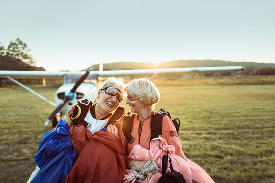 A senior lesbian couple stands on a field, airplane in the backdrop, beaming with pride and exhilaration after skydiving. Their smiles hint at a bucket list dream fulfilled.