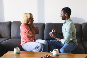 African American man proposing to surprised woman on sofa at home, feeling joyful