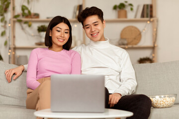 Fototapeta premium Asian Family Couple Watching Movie On Laptop Online Enjoying Evening Together At Home, Sitting On Couch In Cozy Modern Living Room. Weekend Leisure Concept