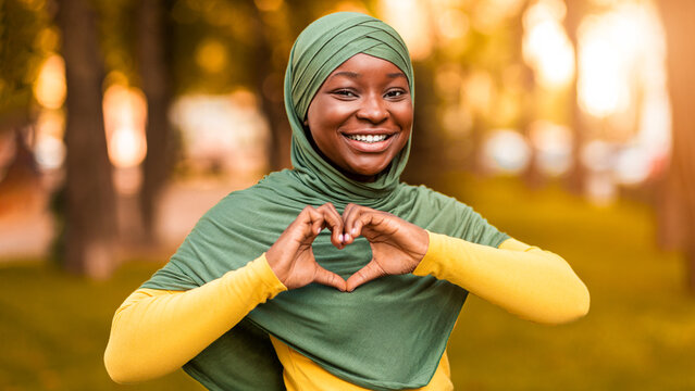 Love Concept. Smiling African Muslim Woman In Hijab Showing Heart Gesture, Holding Hands Near Chest And Smiling At Camera, Positive Black Islamic Lady In Headscarf Posing Outdoors, Free Space
