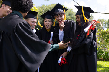 Graduates in caps and gowns celebrating outdoors, excitedly looking at smartphone