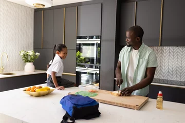 Selbstklebende Fototapeten African American father preparing lunch in kitchen with daughter watching happily © WavebreakMediaMicro