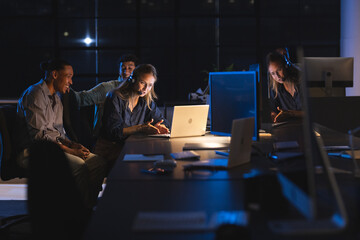 Team collaborating on project at night in modern office, using laptops and headsets
