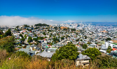 Wide-angle view of San Francisco's residential neighborhoods and downtown skyline, with coastal fog rolling in from the west beneath a clear summer sky.
