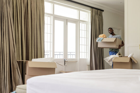 Young African American man carrying moving box with teddy bear and blanket in bedroom, copy space