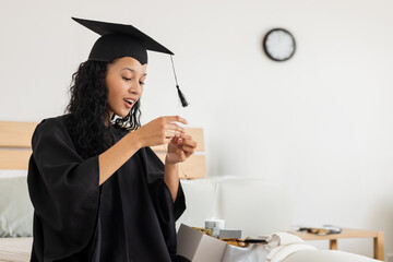 Teenage girl opening gift box wearing graduation gown with mortarboard on bed, examining necklace