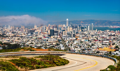 Obraz premium Panoramic view of downtown San Francisco from Twin Peaks, featuring iconic skyscrapers, urban density, coastal fog, and the curving summit roadway under a clear summer sky.