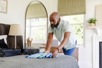 Senior African American man folding striped shirt into black suitcase in bedroom, copy space