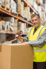 Man in his 50s sealing cardboard box with tape dispenser in warehouse aisle, copy space