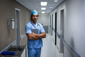 Standing male doctor crossing arms in hospital corridor, holding digital tablet with stethoscope