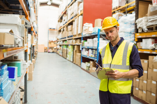 Male warehouse worker inspecting inventory in warehouse aisle, with clipboard and pen, copy space
