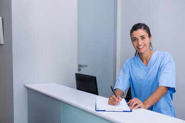 Writing female nurse standing at reception desk in clinic, holding clipboard and pen, copy space