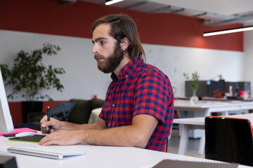 Sketching bearded man using stylus on graphic tablet at open-plan office desk, with sticky notes