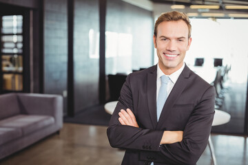 Standing middle-aged man wearing black suit crossing arms in open-plan office, glass partitions