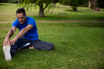 Stretching man grasping extended leg on grass in public park, with running shoes, bench, copy space