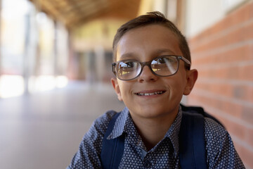 School-age student standing near brick wall in covered corridor, wearing glasses and backpack
