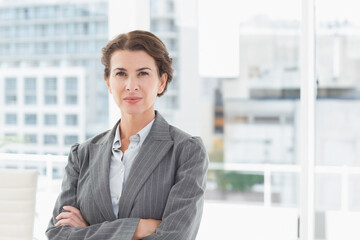 Standing woman crossing arms in modern office, wearing gray pinstriped blazer and light blue blouse