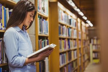 Student is leaning against bookshelves reading hardcover book in library aisle under lights