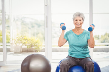 Smiling senior woman lifting blue dumbbells in home gym, with stability ball and glass windows