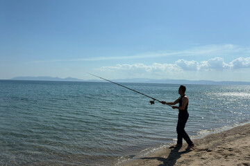 Man fishing on beach at sunset, catching fish rod into calm sea or ocean, outdoor adventure,...