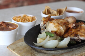 This image features a sizzling plate of grilled chicken accompanied by sides like beans, rice, and...