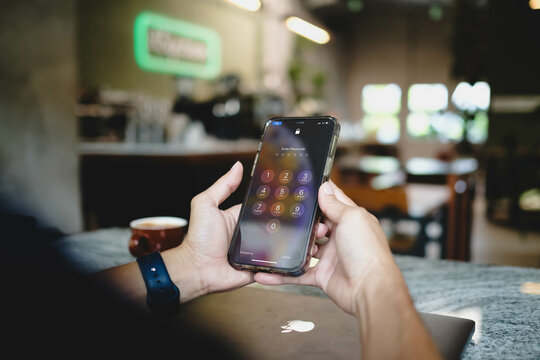 Bangkok, Thailand. May 14, 2025; Person holding smartphone displaying iPhone passcode lock screen in cozy cafe with coffee cup and laptop nearby on marble table.