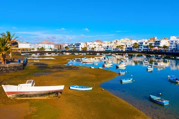 Photo sur Plexiglas Waterfront scene in Arrecife, Lanzarote, with numerous small boats scattered across a shallow lagoon under blue sky. White buildings line background, contrasting with the muddy shore and palm trees