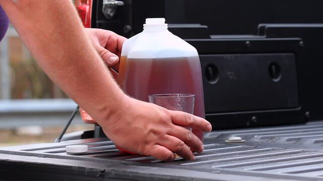 Tea Poured Into A Plastic CupFrom A Gallon Jug On A Truck Tailgate By Male Hands
