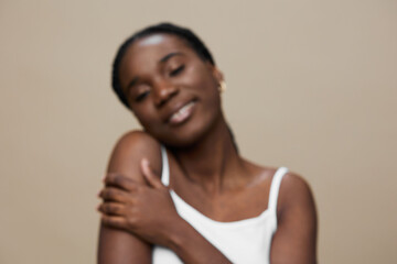 Young African woman smiling gently in a white tank top, embracing herself against a neutral beige...