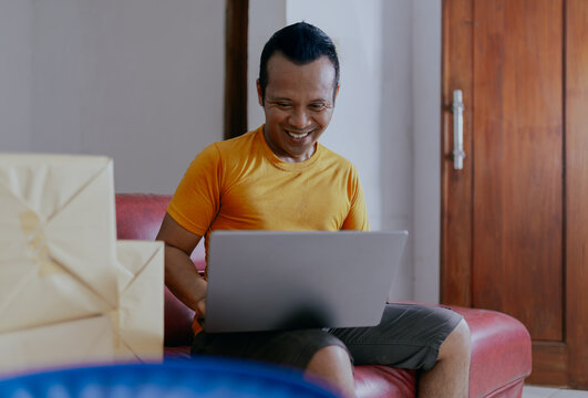 Smiling asian businessman analyzing documents and using laptop while working in home office. Indonesian male sitting on the couch, managing parcel small business remotely, copy space for text.