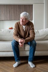 Thoughtful lonely senior man sitting at home with emotional distress. Elderly male on sofa looking around, social abandonment, early dementia, chronic illnesses, mental health issues, life after 60s