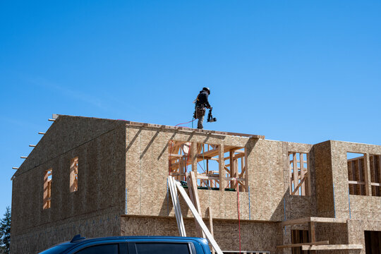 Framing contractor walking across roof of new home construction with a nail gun, sunny blue sky as background, workman on job site
