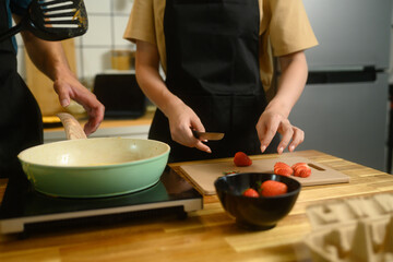 Close up of couple preparing pancakes, slicing strawberries and cooking on stovetop