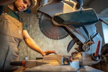 Focused carpenter man in earmuffs use miter saw cutting wooden plank in workshop. Handyman precision cutting, woodworking craftsmanship, blade control, furniture making business, construction hobby