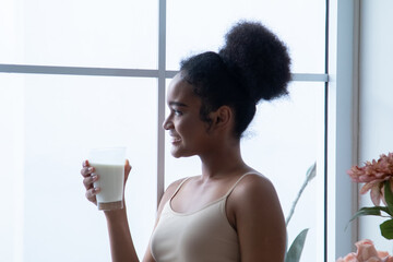 Beauty African woman with healthy lifestyle smiles, holding a fresh milk glass by a bright window;...