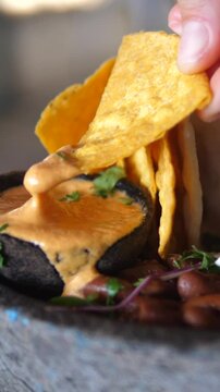 Vertical screen multiple shots of tortilla chips being dipped into a creamy cheese sauce, served alongside kidney beans and garnished with herbs