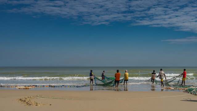 A group of people is pulling fishing nets out of the ocean. Waves are foaming on a sandy beach. Reflection on wet, smooth sand. Blue sky, clouds. Copy space. Madagascar. Morondava. Mozambique Channel.