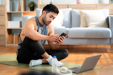Young asian man sitting on yoga mat relax after home exercise using smartphone, modern healthy and digital lifestyle, fitness routine, man looking smart phone after workout, self-care and wellness.