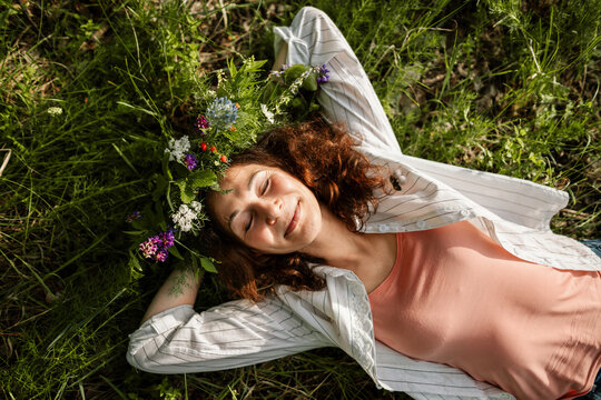Woman lying in grass with flower wreath, eyes closed, enjoying summer nature