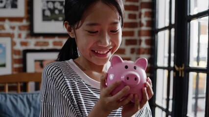 A young girl joyfully holds a pink piggy bank in a cozy room. Captured from a frontal angle, the video conveys a sense of savings and happiness.